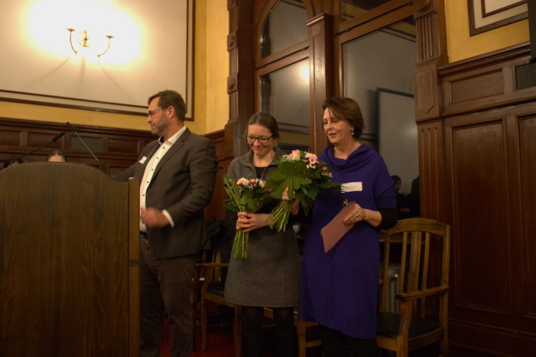 Lins oben eine zweistrahlige Wandlampe. Daruner ein hölzernes Rednerpult. Rechts daneben ein Mann mit weißem Hemd neben zwei Frauen mit Blumensträußen.