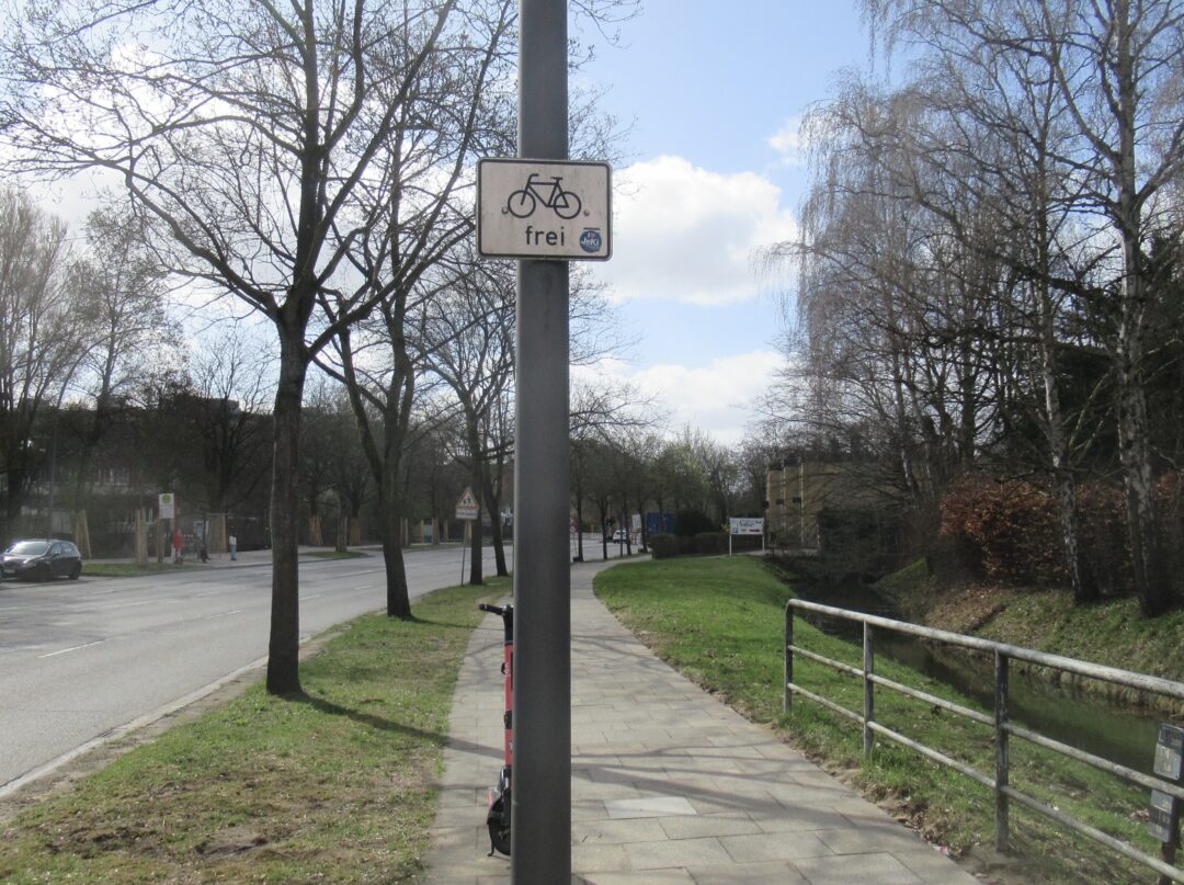 Blick auf den zugewachsenen Radweg., rechts der Gehweg, in der Mutte an einem Pfahl das Zusatzzeichen