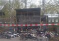 Die abgebrannte Solibox am 13.4.26. In der leeren Blechhülle und davor liegen verbrannte Gegenstände und Kleidungsstücke. Polizei-Absperrband zieht sich im Vordergrund quer durchs Bild. Foto Janwillem van den Loo.