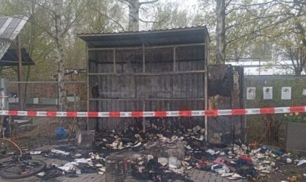 Die abgebrannte Solibox am 13.4.26. In der leeren Blechhülle und davor liegen verbrannte Gegenstände und Kleidungsstücke. Polizei-Absperrband zieht sich im Vordergrund quer durchs Bild. Foto Janwillem van den Loo.