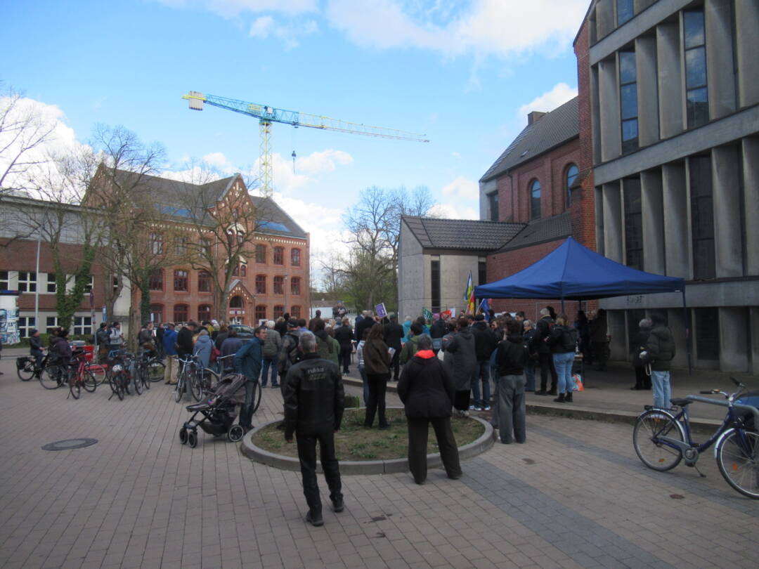 Menschen sehen auf einem Platz. Im Hintergrund ein Schulgebäude. Rechts ein blauer Pavillon vor einer Kirche.