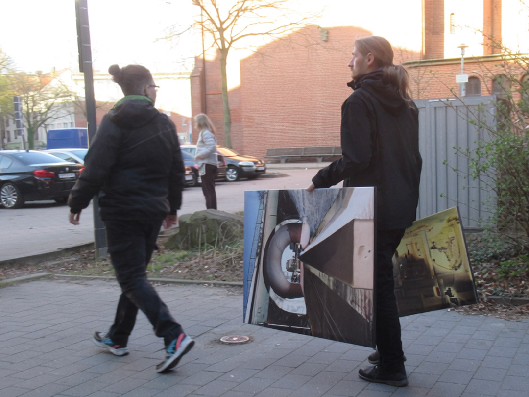 Ein Mann und eine Frau in dunkler Kleidung gehen auf einem grauen Plattenweg. Der Mann trägt zwei Bilder. Im Hintergrund noch eine Frau vor geparkten Autos.