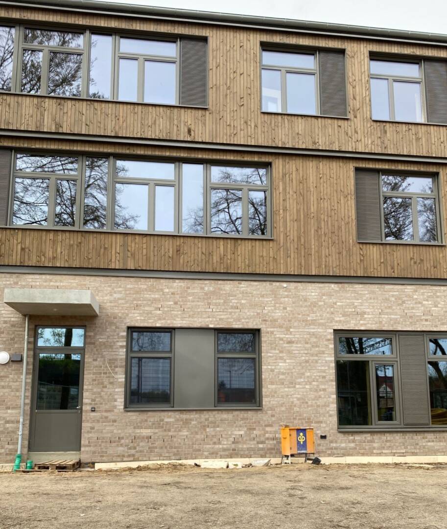 Ein Haus, im Erdgeschoss aus hellem Sandstein mit einer dunklen Tür mit Vordach, daneben zwei Fenster und rechts ein bodentiefes Fenster. Darüber zwei Stockwerke mit Holzverkleidung und großen Fenstern.