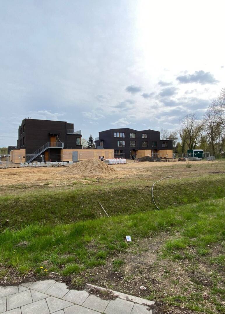 Im Vordergrund eine Wiese und quer ein Graben. darüber ein Sandhaufen. Im Hintergrund dunkle Mehrfamilienhäuser mit hellen Schuppen davor.