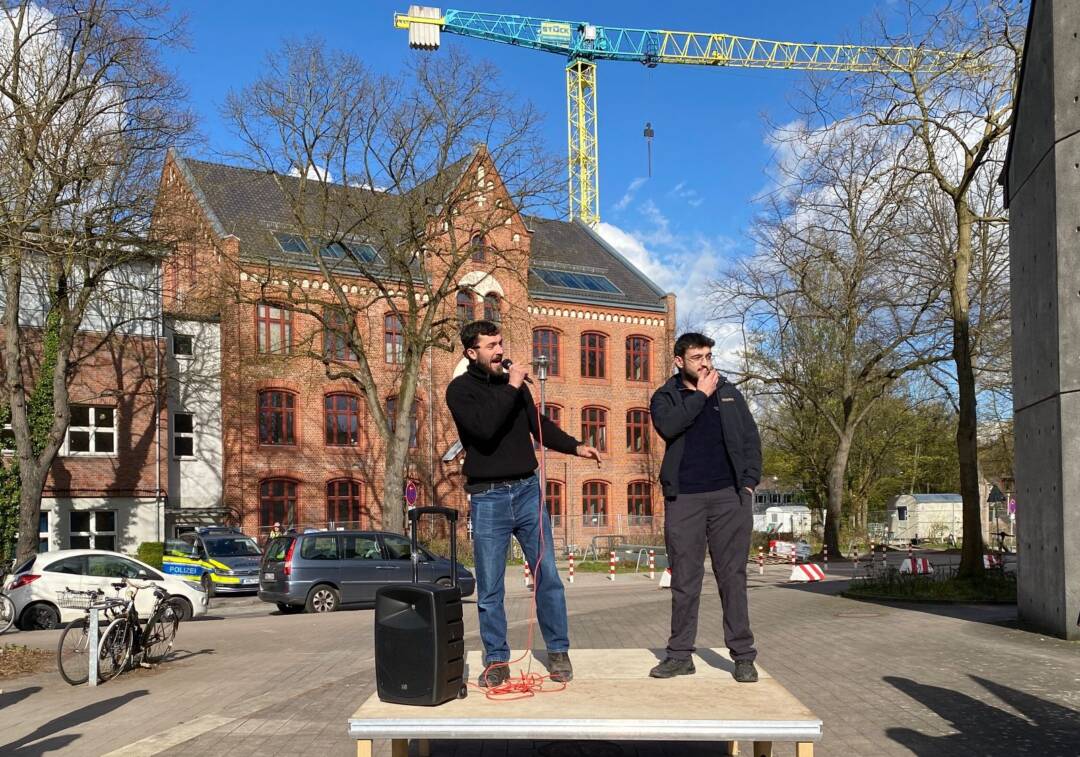 Auf einem hellen Holzpodest stehen zwei Männer in dunklen Pullovern. Im Hintergrund ein Klinker-Schulgebäude. Dahinter ein Kran.