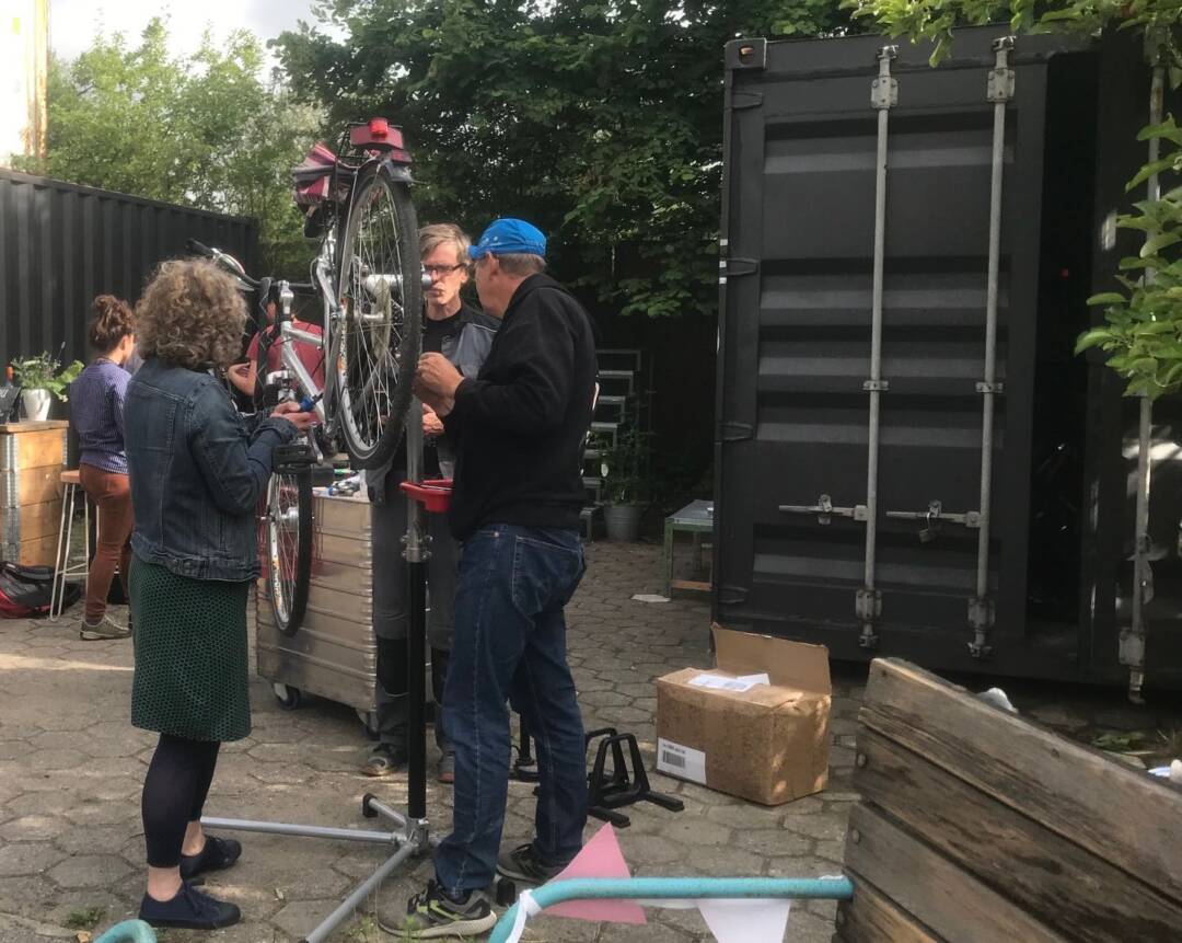 Ein Fahrrad auf dem Kopf steht zwischen einer Frau und einem Mann vor einem Container.