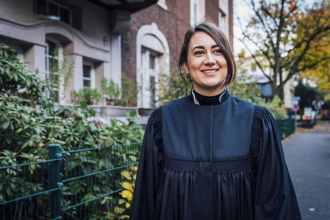 Pastorin Assif im Talar vor einem Haus