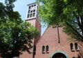 Ein Kirchturm mit drei weissen oberen Etagen neben einem Kirchengiebel aus Backstein. links unten und rechts oben grünes Laub.
