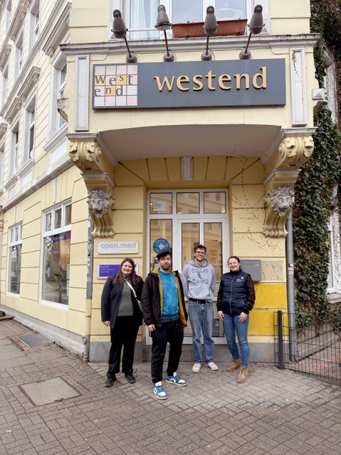 Vor der Ecke eines gelben Gründerhauses, unter dem grauen Schild westend,. stehen vier Menschen. Außen zwei Frauen und in der Mitte zwei Männer.