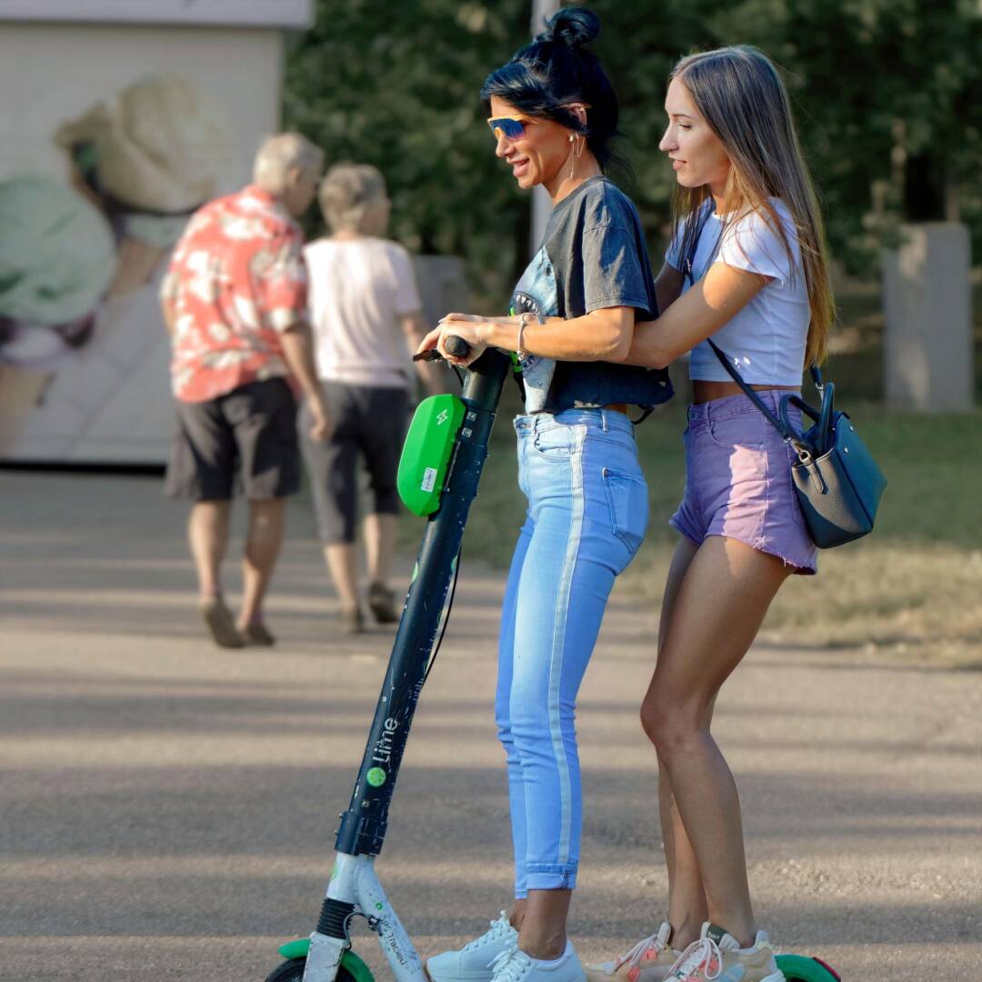 Zwei junge Frauen zusammen auf einem E-Scooter. Die Erste trägt eine blaue lange Hose, die zweite einen Shorts. Im Hintergrund ist noch ein Paar in kurzen Hosen.