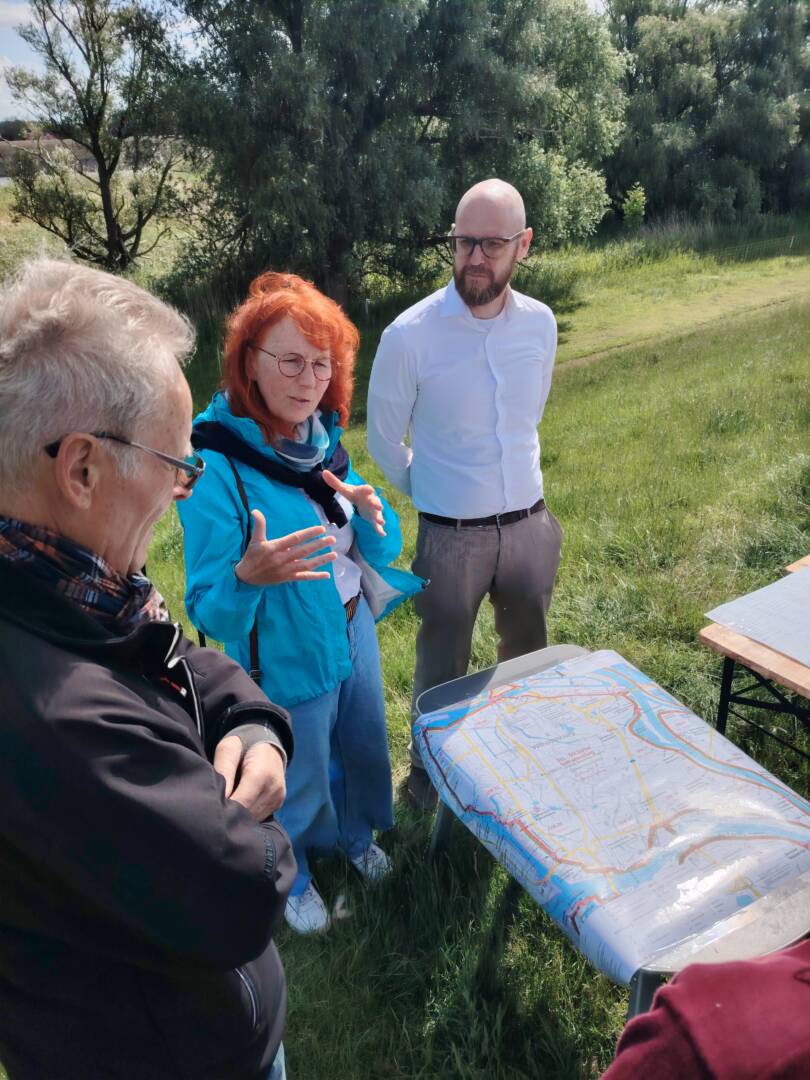 Links ein Mann in schwarzem Anorak, rechts daneben eine Frau mit roten Haaren im türkisfarbenen Regenanzug und hinten in der Mitte ein Mann mit Glatze, dunklem Bart in weißem Hemd. Davor eine Landkarte von Wilhelmsburg.
