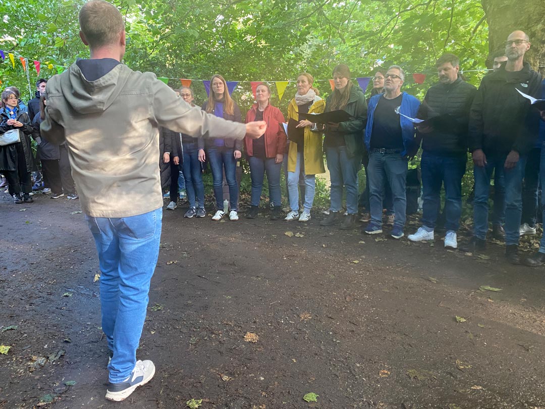 Im Vordergrund der Chorleiter in Jeans und heller Jacke. Im Hintergrund, aufgereit am Wegesrand der gemischte Chor in wetterfester Alltagskleidung.