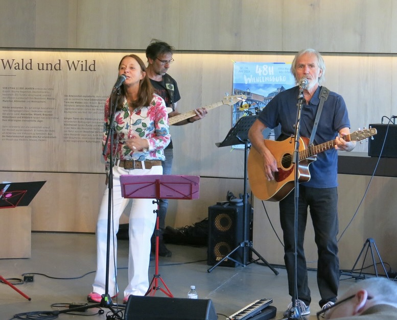 eine singende Frau in bunter Bluse und weißer Hose, ein Mann in Stoffhose/Shirt mit Akkustikgitarre. Im Hintergrund ein Bassist, der von der Frau verdeckt wird. Foto: Hermann Kahle