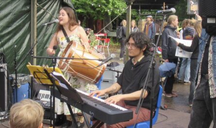 Im Vordergrund ein Keyboarder mit dunklem Kapuzenpulli an seinem Instrument. Im Hintergrund eine Frau im Sommerkleid, die in ein Mikrofon singt und eine große Trommel aus Holz festhält und schlägt. Foto: Hermann Kahle