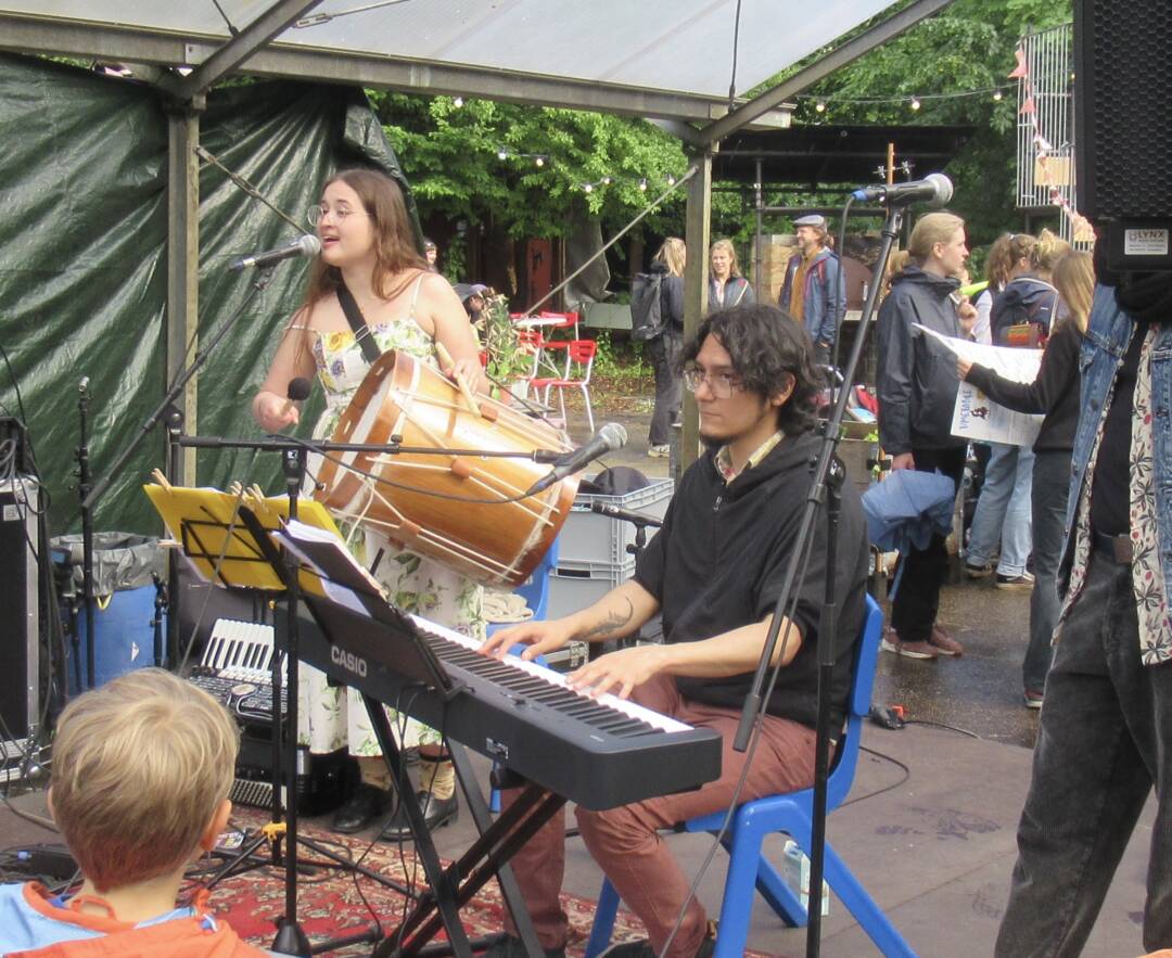 Im Vordergrund ein Keyboarder mit dunklem Kapuzenpulli an seinem Instrument. Im Hintergrund eine Frau im Sommerkleid, die in ein Mikrofon singt und eine große Trommel aus Holz festhält und schlägt. Foto: Hermann Kahle