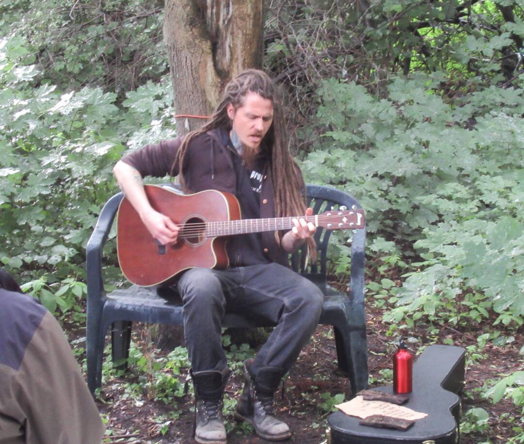 Ein Gitarrist mit Dreadlocks und Bart in robuster Freizeitbekleidung sitzt im Wald auf einer Plastikbank. Foto: Hermann Kahle