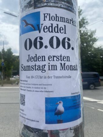 Ankündigungsplakat für den Flohmarkt. Es klebt an einem Laternenpfahl. Im Hintergrund sieht man eine Straße.