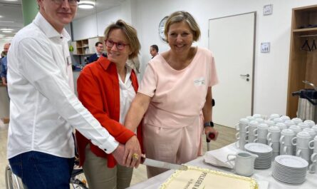 Ein Mann und zwei Frauen stehen hinter einer Marzipantorte mit einem Kuchenmesser in den drei Händen, die sich anfassen.