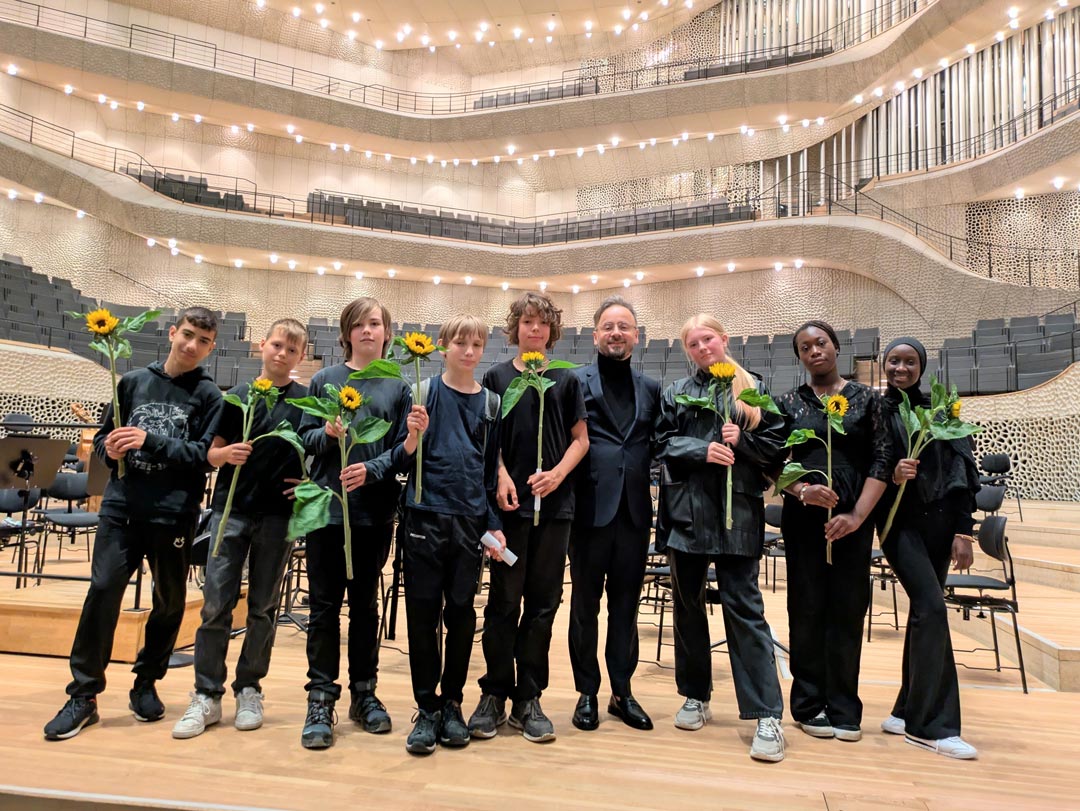8 schwarz gekleidete Schüler*innen auf der Bühne der Elbphilharmonie. Ihren Lehrer haben sie in die Mitte genommen. Alle außer dem Lehrer halten eine Sonnenblume. Alle sehen zufrieden aus. Foto: Iris Hahn-Möller