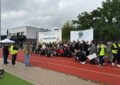 Auf einem Sportplatz mit rotem Untergrund sitzen, liegen stehen alle Beteiligten des Cups in einem großen Halbkreis vor zwei Bannern auf denen der Name des Turniers und das Logo des IJB abgebildet sind. Davor stehen drei Männer, einer hält eine Ansprache. Foto: Necmettin Bozkurt