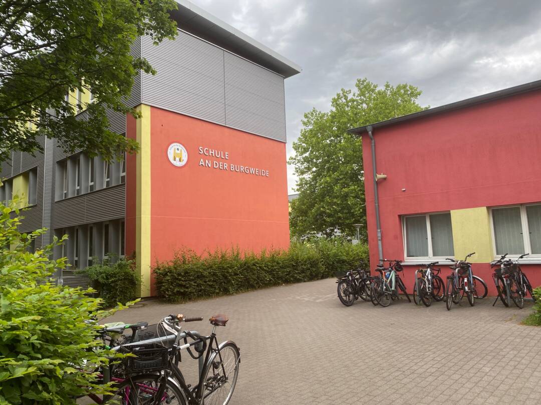 Links unten ein Gebüsch, daneben ein Fahrrad. Darüber ein hohes Gebäude, das obere Drittel grau. darunter ziegelrot. Die vordere Ecke ist gelb. Daneben ein großer Busch und davor das nächste Gebäude in ziegelrot mit zwei Fenstern, die durch einen gelben Streifen getrennt sind. Davor vier Fahrräder. Dazwischen ein Asphaltweg.