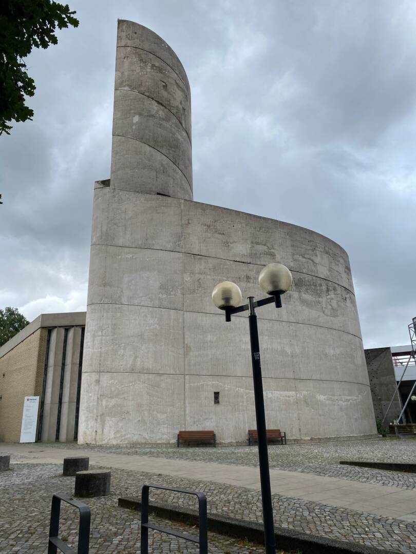 Vor einem grauen Himmel steht eine Betonkirche mit einem in die Höhe gewendelten Turm. Davor steht eine Straßenlampe mit zwei Glaskugel-Lampen.