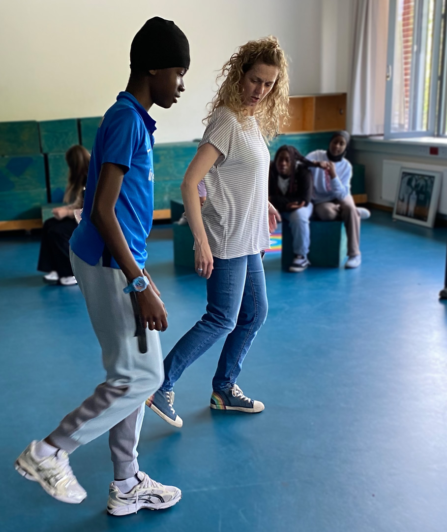 Im Vordergrund zeigt die Pädagogin dem Schüler Tanzschritte. Im Hintergrund sitzen andere Schüler*innen und gucken zu. Foto: Iris Hahn-Möller