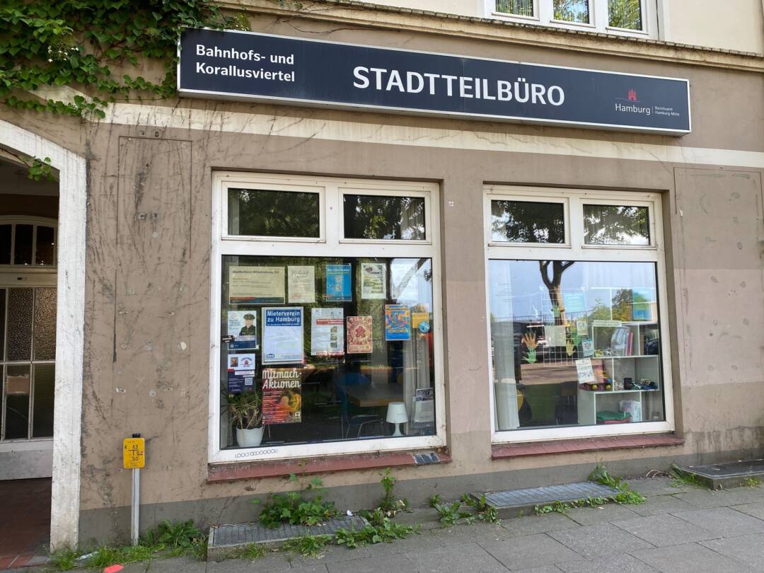Über zwei großen Schaufenstern mit Plakaten ist ein blaues Schild mit weißer Schrift: Bahnhofs- und Korallusviertel Stastteilbüro. Rechts auf dem Schild ist das Wappen von Hamburg. Links im Bild Teil eines Eingangs. Darüber grünes Laub.
