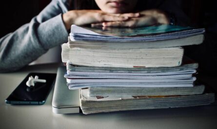 Ein Stapel Bücher Hefte. Darauf hat jemand (wahrscheinlich ein Teenager) Arme und Kinn abgelegt. Unter dem Stapel guckt die Ecke eines Laptops hervor. Daneben liegt ein Smartphone mit Kabellosen Kopfhörern. Düstere Stimmung. Foto: Sweetlouise/Luisella Planeta LOVE PEACE auf Pixabay