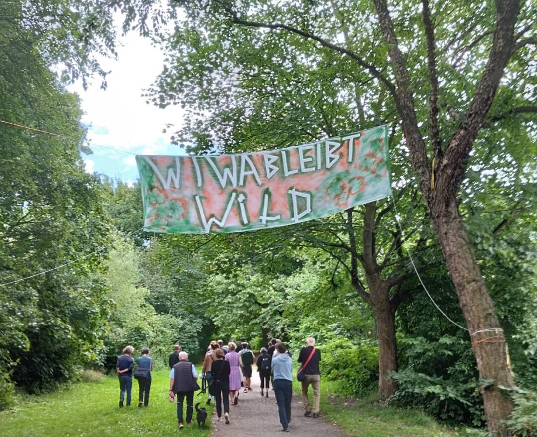 Menschen auf einem schönen Waldweg, darüber ist ein banner gespannt. Darauf steht: WiWa bleibt wild.