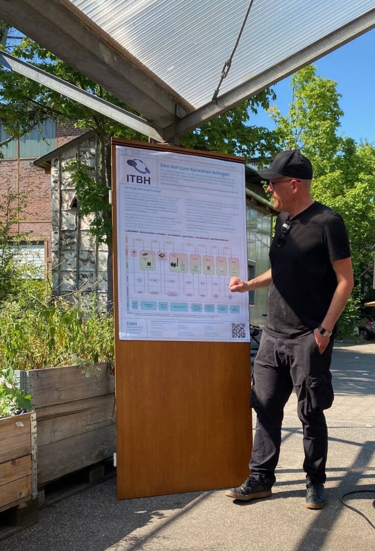 Ein Mann in schwarzem T-Shirt, Jeans und Käppi steht vor einer hölzernen Infowand.
