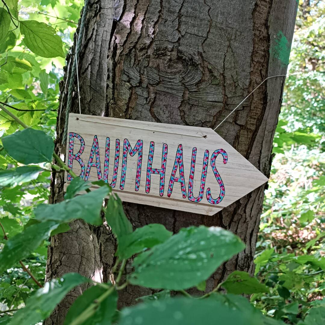 An einem Baumstamm hängt ein Holzschild in Pfeilform. darauf steht in schöner Schrift: Baumhaus.