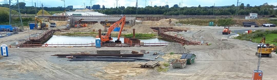 Eine Straßenbaustelle. in der Mitte ein quadratisches Wasserbecken. Davor ein Bagger.