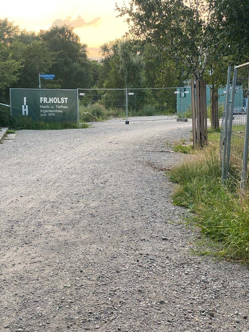 Vorne ein Schotterweg. Dahinter ein Baustellenzaun mit einem Firmenschild.