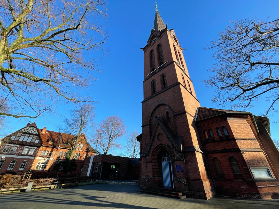 Ein künstlerisches Foto mit stark „stürzenden Linien“. Im Zentrum steht der Turm von St. Emmaus. Der Himmel ist strahlend blau, die Bäume links und rechts haben keine Blätter (Winter). Foto: Lars Meyer