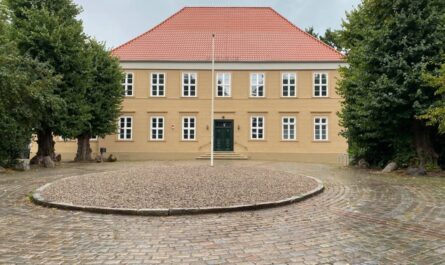 Hinter einem großen Pflastersteinplatz steht ein ockerfarbenes zweistöckiges Gebäude mit rotem Ziegeldach und weißen Sprossenfenstern.