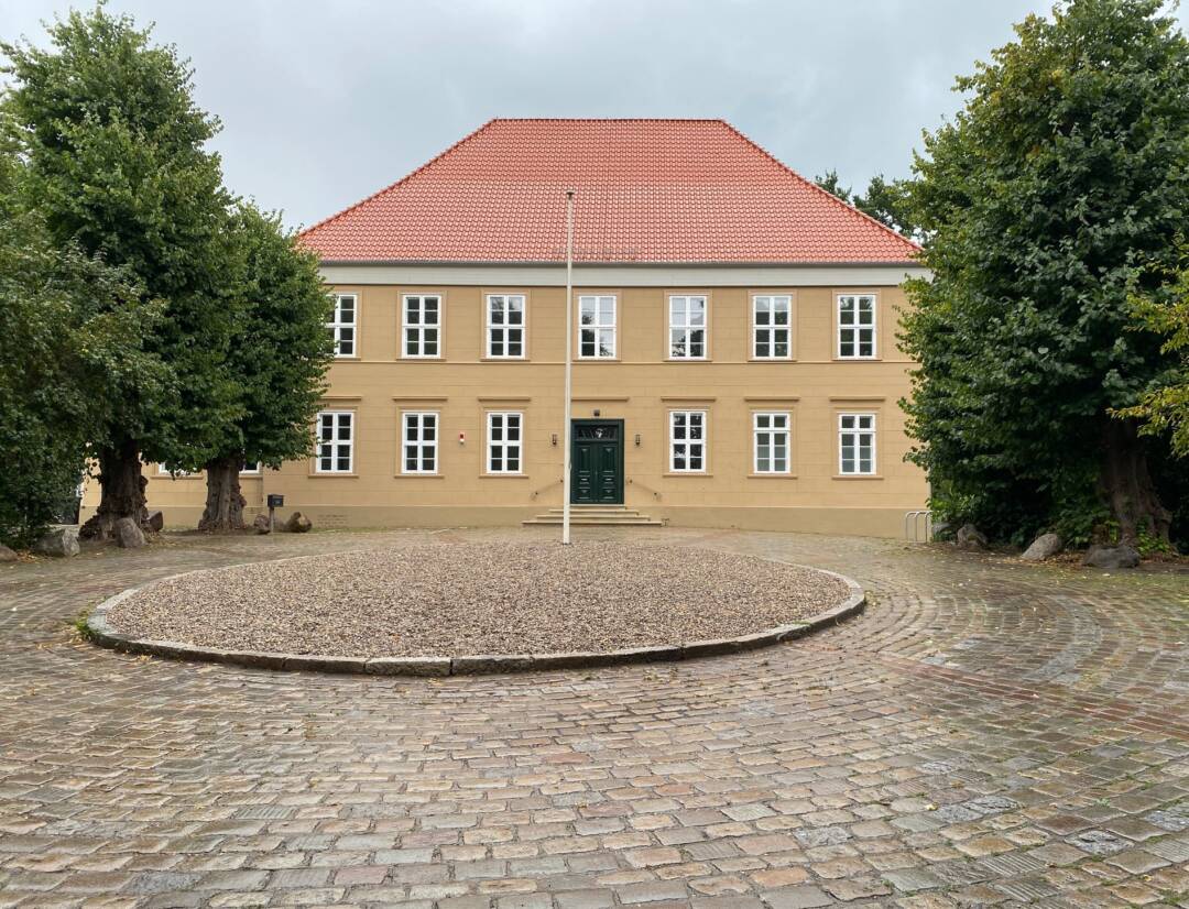 Hinter einem großen Pflastersteinplatz steht ein ockerfarbenes zweistöckiges Gebäude mit rotem Ziegeldach und weißen Sprossenfenstern.