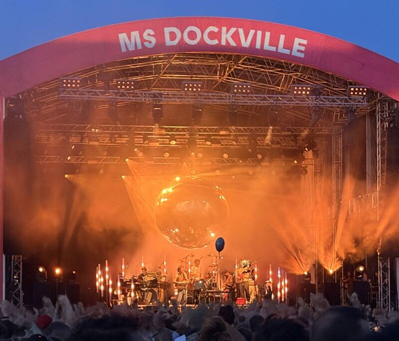 Große Bühne mit einer riesigen Spiegelkugel, die über der Band hängt. Das Licht auf der Bühne ist golden und bildet einen schönen Kontrast zum knallblauen Himmel des späten Nachmittags. Vor der Bühne Fans mit erhobenen Armen (sie feiern).