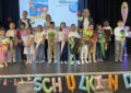14 Kinder und 3 erwachsene Lehr-/Fachkräfte auf einer Bühne. Die kinder halten Schultüten und sind fröhlich.