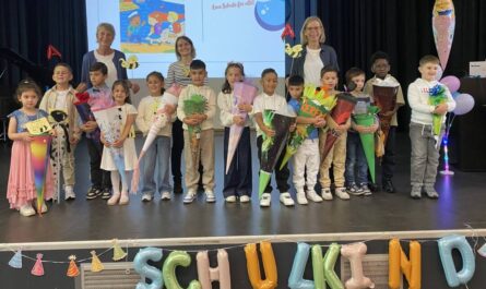 14 Kinder und 3 erwachsene Lehr-/Fachkräfte auf einer Bühne. Die kinder halten Schultüten und sind fröhlich.