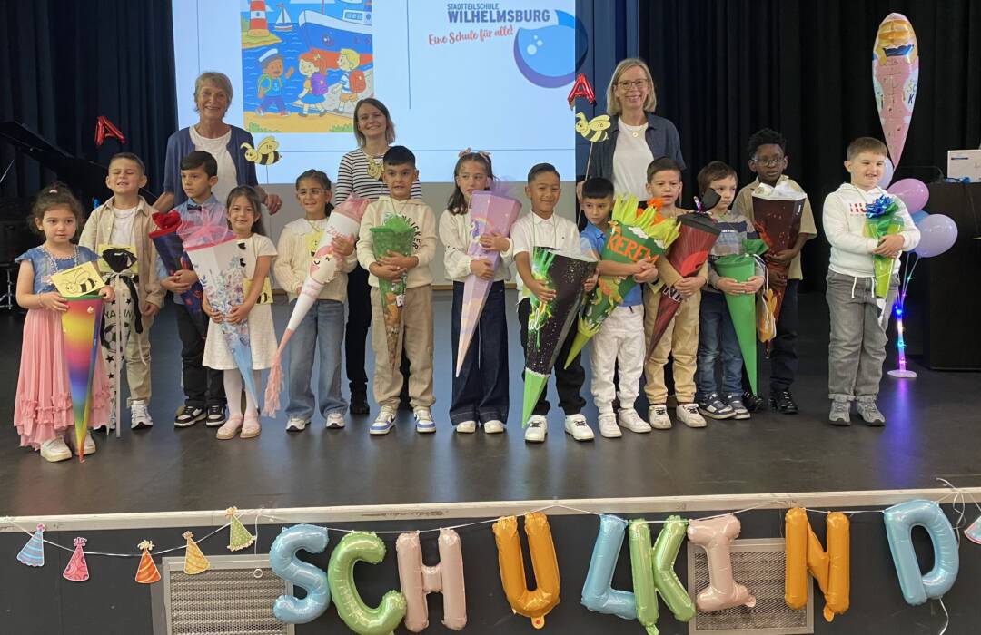 14 Kinder und 3 erwachsene Lehr-/Fachkräfte auf einer Bühne. Die kinder halten Schultüten und sind fröhlich.