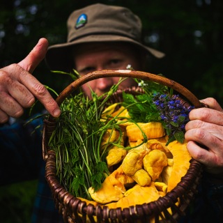 Ein Mann mit einem Filzhut hält einen Korb voller Pilze und Kräuter in die Kamera.