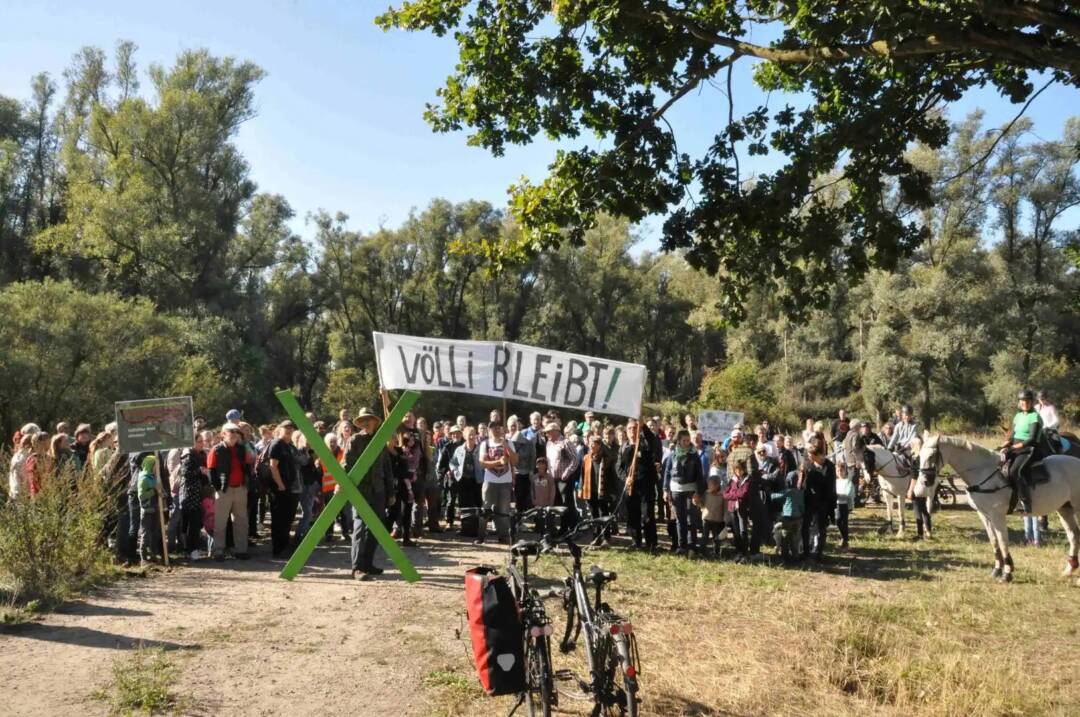 Menschen haben sich auf einer Wiese an einem Waldrand versammelt. Auch berittene Polizei ist zu sehen. Die Gruppe hält ein Banner hoch, darauf steht: Völli bleibt! Außerdem halten sie ein großes grünes X vor sich.