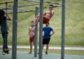 Drei männlich und eine weiblich gelesene Person am und um ein Outdoorfitnessgerät. Im Hintergrund ein Hang/Deich mit Gras Bild von Edmund_H auf Pixabay