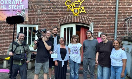 Vor dem Cafe Nova, das in einem alten, urbanen Backsteingebäude untergebracht ist, stehen zwei Männer mit Kamera- und Ton-Equipment, sowie 6 weitere Personen. Alle strahlen in die Kamera