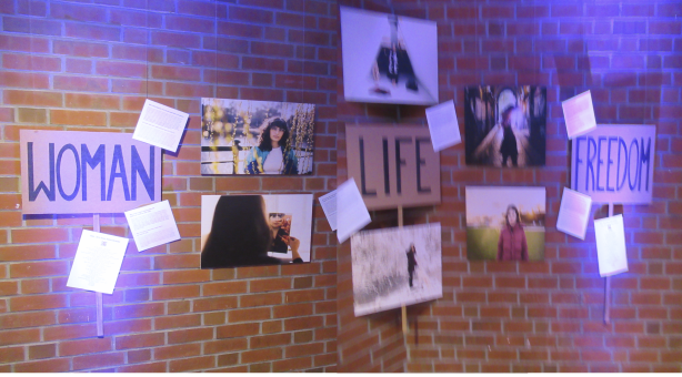 Fotoausstellung am dewr Mauer mit mehreren Potraitfotos von Frauen und Zetteln mit Texten. Auf drei Schrifttafeln üner die AUsstellung verteilt die Wörter Woman, Life, Freedom