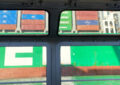Blick aus einer Bakasse: Im Vordergrund die Bordwand mit den Fenstern, dahinter eine riesige Bordwand (grün) und oben auf dem Schiff zig Container. Die Sonne scheint. Foto: Ania Groß