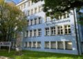 Ein hellblaues Schulgebäude mit fonf Reihen Fenstern. Links unten und rechts oben Teil von Bäumen.