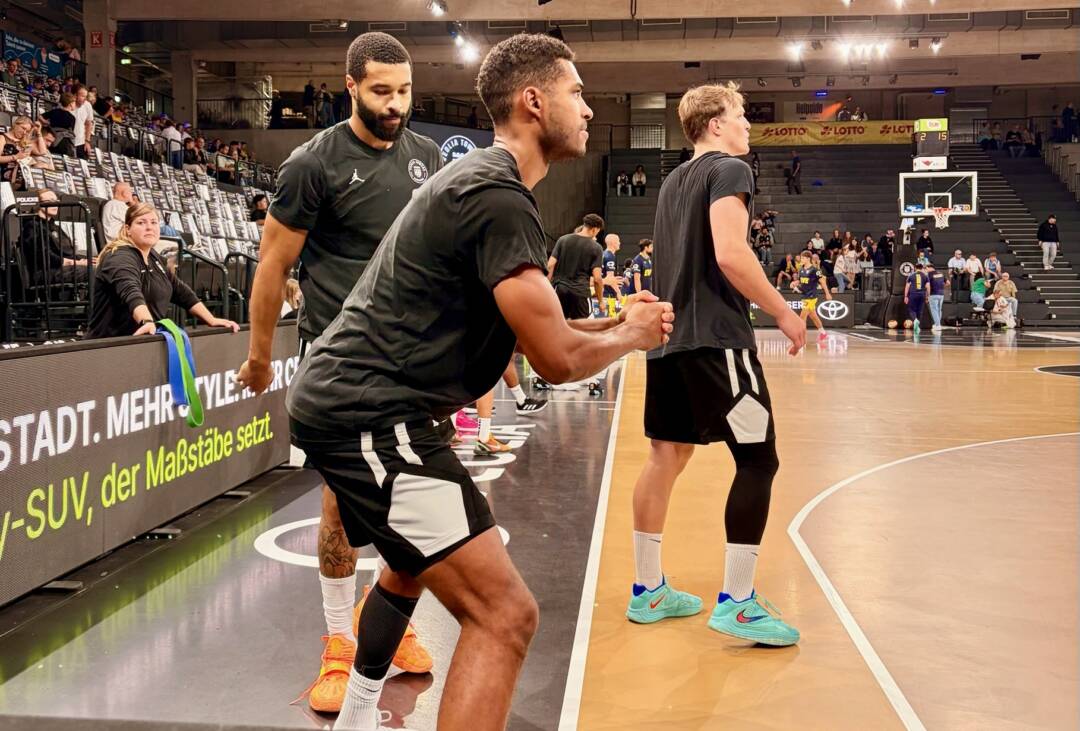 Im Vordergrund drei junge Männer in schwarzen Sportklamotten, die verschiedene Bewegungen machen. Im Hintergrund die Halle. Es sind noch wenige Zuschauer*innen auf den Rängen.