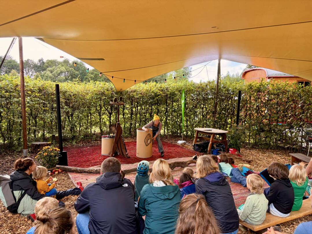 Unter einem gelben Sonnen-/Regensegel spielt eine erwachsene Person Theater und viele Kinder sitzen davor und gucken gebannt in die Richtung